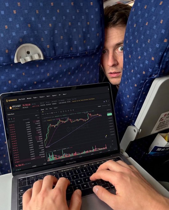 A man squeezes between airplane seats while intently trading Bitcoin on Binance via a MacBook Pro, with the BTC/USDT chart showing 78,764.50—proving that even with a dead phone, market watching waits for no one.