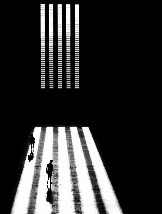A dramatic black and white image of two silhouetted figures walking down a corridor illuminated by a striped pattern of light shining from a slatted window above.