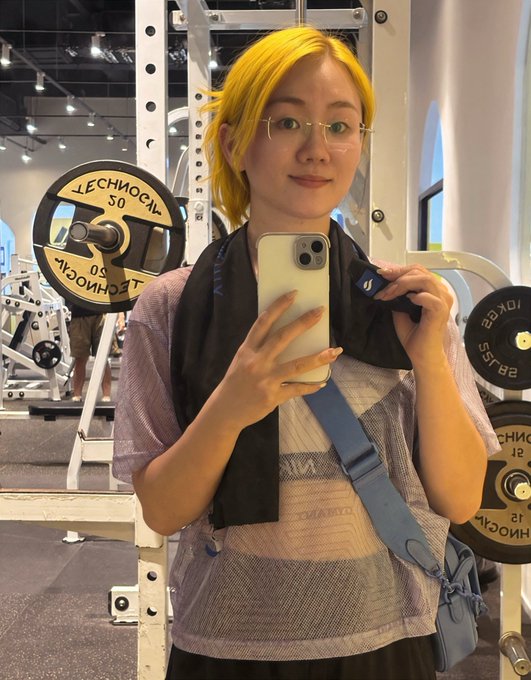 A person with bright yellow hair and glasses takes a mirror selfie with a smartphone inside a gym, with weight plates visible in the background.