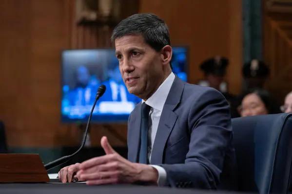 A man in a dark blue suit and tie speaking into a microphone at what appears to be a congressional or senate hearing, gesturing with his hands.