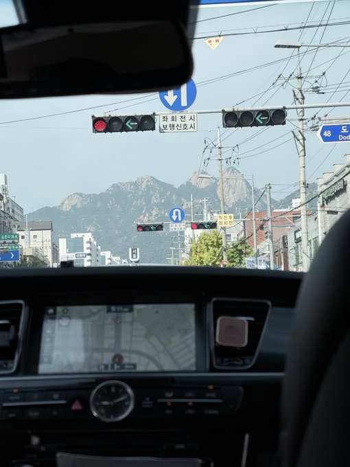 The image, taken from inside a car on a road in South Korea, shows traffic lights and a GPS navigation screen while the dramatic rocky peaks of Bukhansan mountain rise in the distance, with a Korean road sign indicating the direction to "북한산로" (Bukhansan-ro). This perfectly sets the scene for the post about running uphill—Bukhansan is one of Seoul's most iconic hiking destinations, suggesting the driver is heading toward the trails for a run with @kbwofficial.
