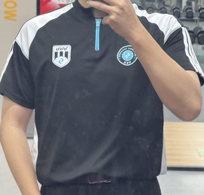 A man takes a mirror selfie wearing a black athletic shirt with white shoulder panels, a blue zipper, and logos including a shield with the number '2' and a 'PROFESSOR JAGUAR' emblem.
