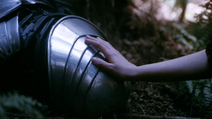 A cinematic close-up of a pale hand reaching out to touch a smooth, metallic, cylindrical object—possibly a helmet or part of a suit—resting among dark green foliage in a dimly lit forest.