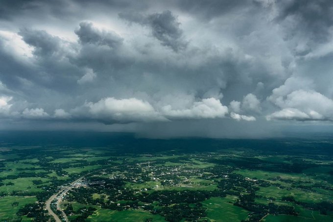 An aerial view shows dark storm clouds looming over a green rural landscape with scattered villages and winding roads.