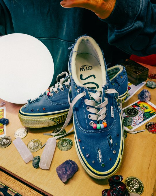 A pair of distressed blue TOMS sneakers decorated with colorful beads and gems sits on a wooden table surrounded by various items including coins, tarot cards, a crystal, seashells, and a person wearing a green sweatshirt gesturing with their hand.