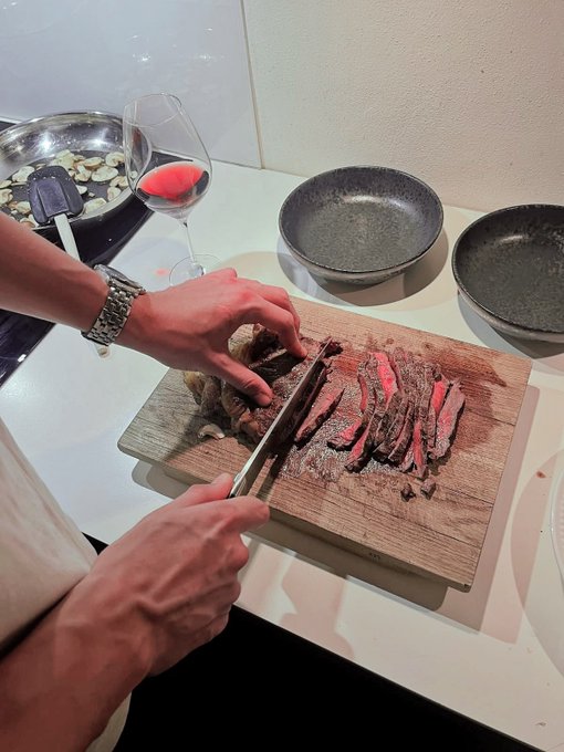 A person wearing a silver watch slices a medium-rare steak on a wooden cutting board, with a glass of red wine and empty plates nearby.