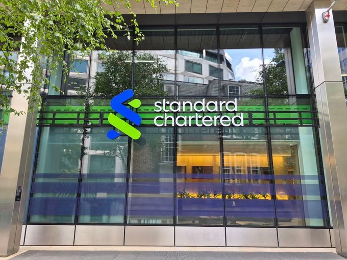 The exterior of a Standard Chartered bank branch, featuring the company's green and blue logo on a modern glass facade.