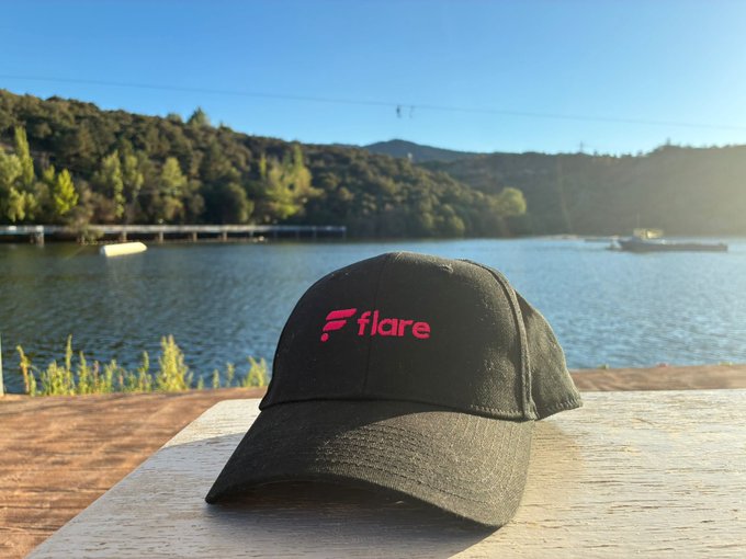A black baseball cap with a pink 'Flare' logo sitting on a wooden surface with a scenic lake and forested hills in the background.