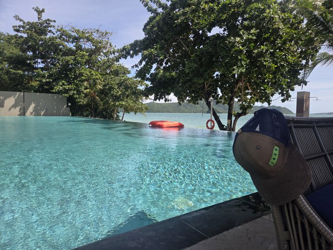 The image features an outdoor swimming pool with a wicker chair placed next to it. A hat is hanging from the edge of the pool, indicating that someone has been there recently. The water appears to be calm, suggesting a peaceful atmosphere. In the background, there are trees and a boat floating on the water, adding to the serene setting. The post about the wicker chair by the swimming pool seems unrelated to this scene, as it does not provide any context or information about the location of the chair or its connection to the image.