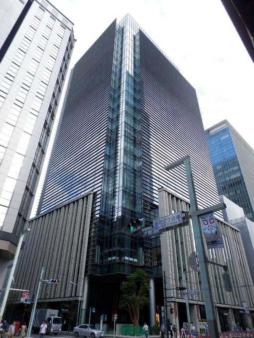The image shows a tall building with many windows, including one that has a sign saying "50". The building appears to be a skyscraper or office building located in Japan. In the background, there are other buildings as well. A street sign is visible on the right side of the photo. There are also people walking around and cars driving by, indicating an active urban environment.

The image includes a post that talks about Bitcoin and cryptocurrency adoption in Japan. The post mentions that SBI Shinsei Bank will allow customers to convert their deposit interest into Bitcoin, Ethereum or Ripple starting from June 10th. This development is aimed at promoting the use of digital assets as part of traditional banking services, which could potentially lead to mainstream acceptance of cryptocurrencies.