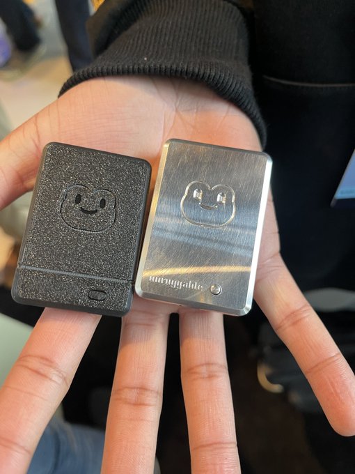 Two compact rectangular metal devices with a smiling face logo, one black and textured, the other silver and shiny, held in a person's hand.