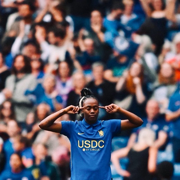 A female soccer player wearing a blue Chelsea jersey with the 'USDC BY CIRCLE' sponsor logo, posing with her fingers to her head in front of a blurred crowd.