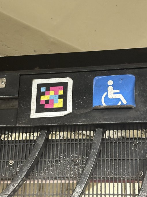 A pixelated sticker with colorful squares is affixed next to a blue wheelchair accessibility sign above a metal grate.