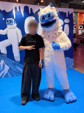A person stands next to a blue and white furry mascot costume on a blue carpet at what appears to be an indoor event or convention.