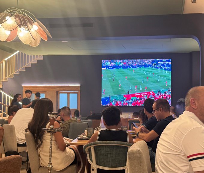 A group of people gathered in a dimly lit lounge watching a soccer match on a large wall-mounted TV, with some holding drinks and a hookah visible in the foreground.