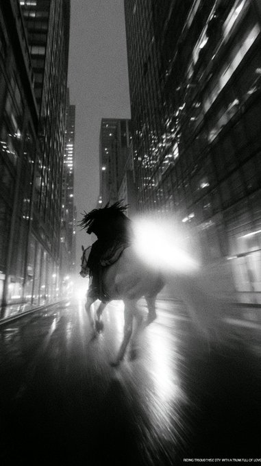 A black-and-white artistic image showing a silhouette of a person riding a horse down a city street at night, with bright lights creating a dramatic glow.