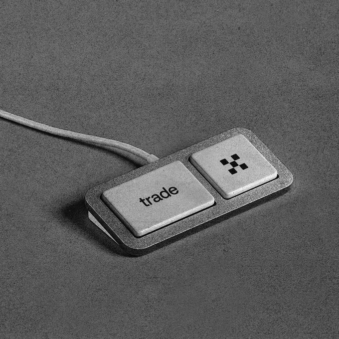 A sleek metallic device with two buttons, one labeled 'trade' and the other featuring a four-dot symbol, connected by a braided cable on a gray surface.
