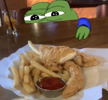 A basket of fried chicken and fries with ketchup on a wooden table, with a cartoon Pepe the Frog peeking over the table and a glass of iced drink in the background.