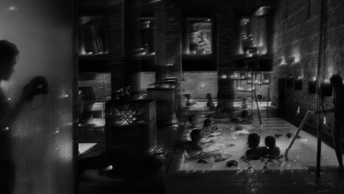 A black and white image of a dimly lit indoor pool area with several people bathing, surrounded by brick walls and ambient lighting from candles and hanging fixtures. The scene captures a moody, atmospheric gathering that reflects the post's tone of enjoying a fun yet anonymous night out, staying in the background while soaking in the warmth and company.