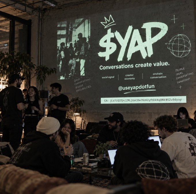 An event space with a large projection displaying '$YAP' and 'Conversations create value' along with '@useyapdotfun' and a crypto contract address, while people socialize and work on laptops.