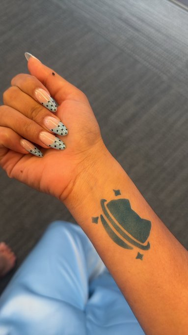 A person displays a wrist with a black Saturn planet tattoo and nails painted with a light blue polka dot design.