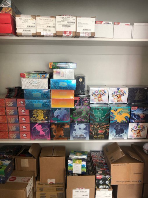 A photograph of a closet or storage shelf packed with various sealed booster boxes and trading card game products, including Pokémon and Yu-Gi-Oh! boxes, with shipping boxes stacked on the top and bottom shelves.