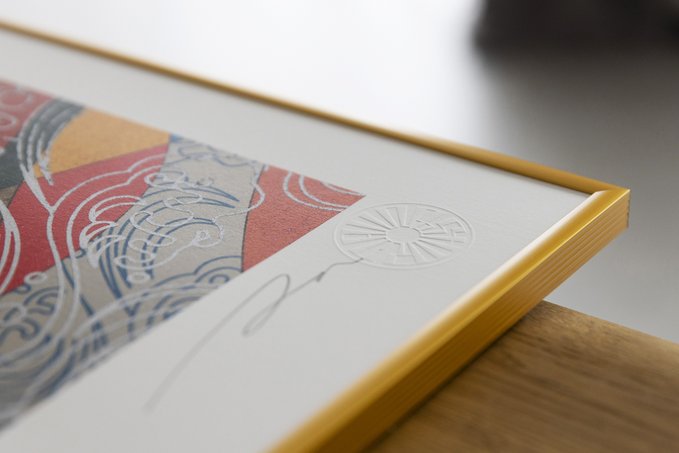 A close-up of a framed artwork featuring colorful abstract designs with gold foil accents and an embossed circular seal in the corner of the white matting.