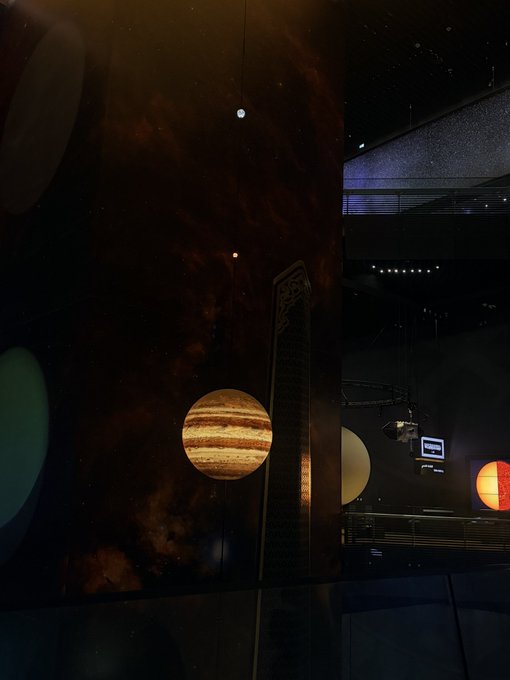 A dimly lit photograph of a museum exhibit displaying suspended models of planets, prominently featuring Jupiter and the Sun against a large cosmic backdrop.