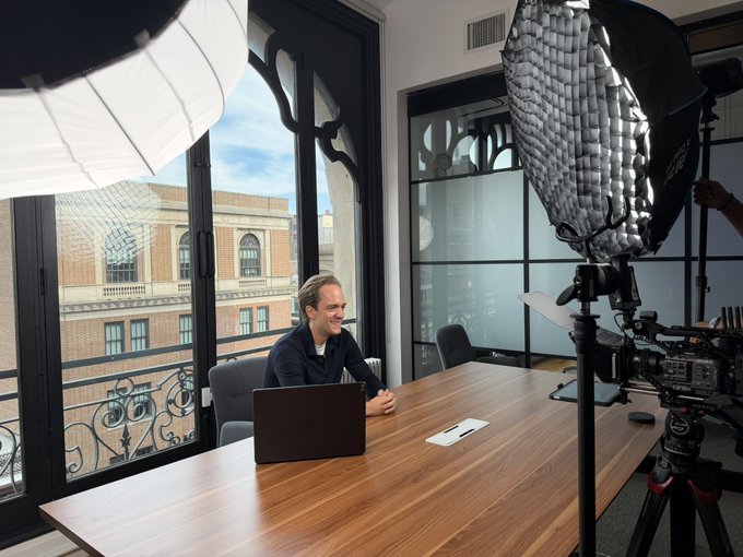 A man sits at a wooden desk with a laptop during what appears to be a video production setup, with professional lighting equipment and a camera pointed at him. The accompanying text suggests this is a filmed "announcement of an announcement" featuring Max Wallenberg, likely teasing a forthcoming business or personal reveal.
