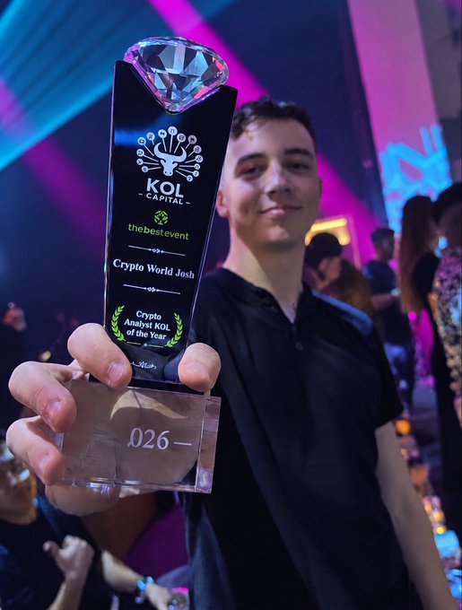 A young man smiles while holding up a crystal trophy featuring a diamond on top, engraved with 'KOL CAPITAL the best event Crypto World Josh Crypto Analyst KOL of the Year' and the number '026'.