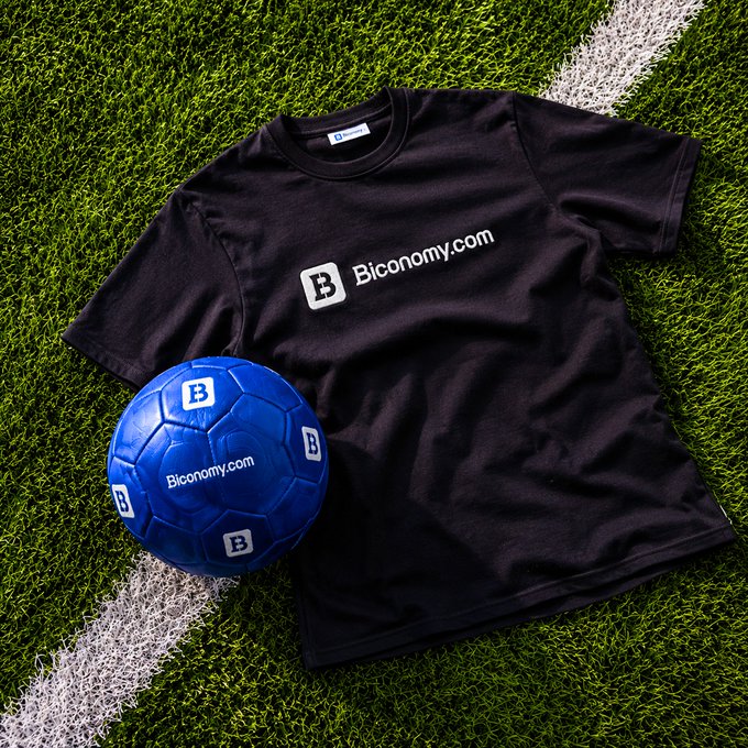 The image features a black t-shirt laying on a green grassy field, with a blue soccer ball next to it. The text "Biconomy.com" can be seen on the t-shirt. This suggests that the person wearing the t-shirt might have an interest in both football and cryptocurrency trading or investing.