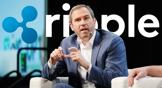 Brad Garlinghouse, CEO of Ripple, speaks while gesturing with his hands at a panel, seated in front of a large backdrop displaying the Ripple logo and name.