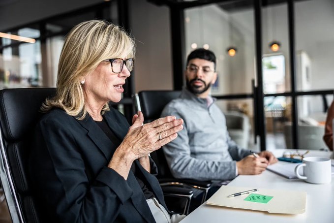 A blonde woman in a black blazer and glasses speaks expressively with her hands clasped in an office meeting, while a bearded man in a gray shirt sits behind her listening.