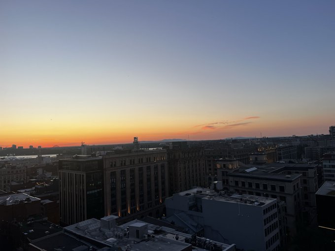 The image shows a city skyline at sunset with buildings of various sizes and designs. The sky has a beautiful orange hue as the sun sets behind the buildings, creating a stunning backdrop for the scene. The photo captures the essence of an urban landscape during this time of day, emphasizing the contrast between the warm colors of the setting sun and the cool tones of the cityscape.