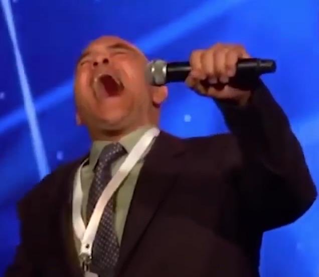 A man in a dark suit, white shirt, and patterned tie passionately singing into a microphone with his eyes closed, set against a blue background with light streaks.