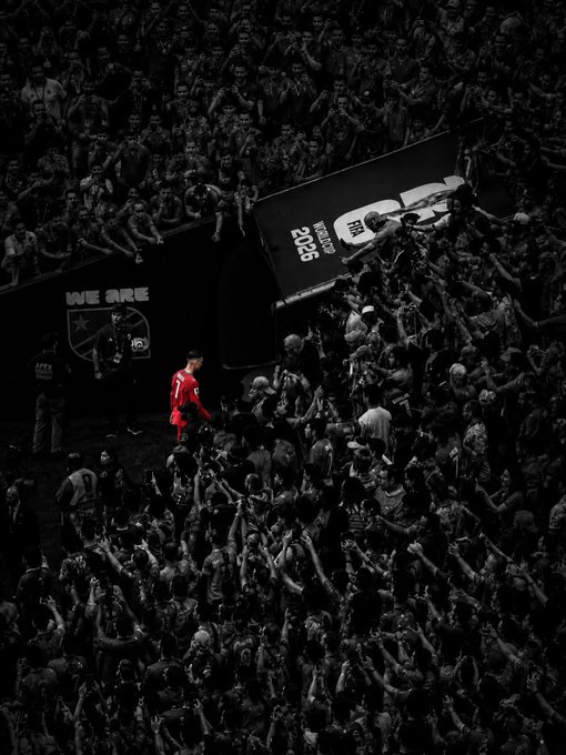 A dramatic black-and-white photo of a soccer player in a red uniform standing alone among a massive crowd of fans, with 'WE ARE' and 'SPORT' signage visible.