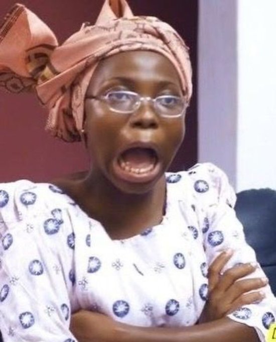 A woman wearing glasses, a pink head wrap, and a patterned white shirt is captured with a shocked expression, mouth wide open.