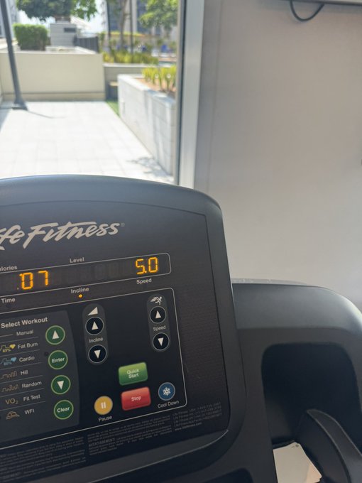 A Life Fitness treadmill console displays workout metrics including an incline of 5.0 and speed of 5.0, with the outdoor view visible through a nearby window.