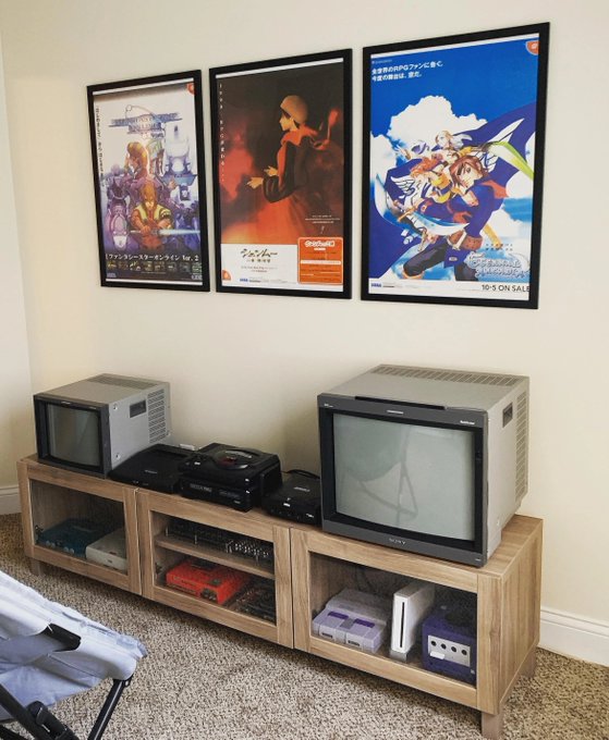 A nostalgic gaming corner featuring three framed anime posters above a wooden entertainment console that houses a vintage CRT television, a small silver monitor, and various retro gaming consoles and cartridges stored on the lower shelves. The posters appear to promote Japanese anime releases, with visible text including "19-3 ON SALE" on the rightmost poster.