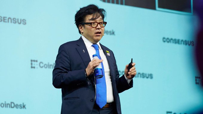 A man in a dark blue suit and blue tie speaking on stage at a CoinDesk Consensus event, holding a water bottle and presentation clicker.