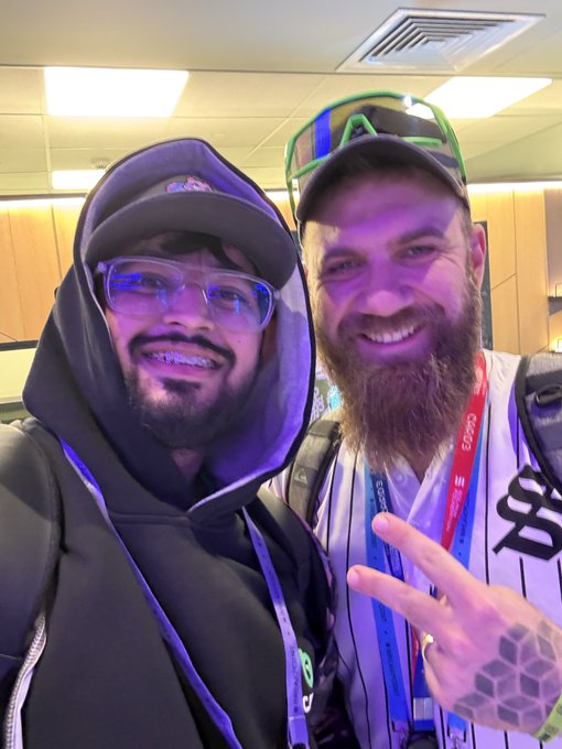 Two smiling men posing together indoors; the right man has a beard, green sunglasses on his cap, and lanyards with "CEGA" text.