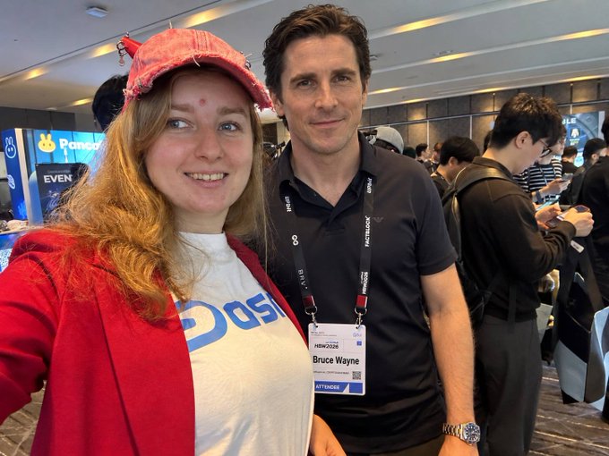 Two people posing for a selfie at an event; the man wears a black polo shirt with a lanyard badge that reads 'Bruce Wayne' and 'ATTENDEES' at 'NEXUS 2026', while the woman wears a red cap and red jacket over a white shirt.