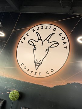 An illuminated circular sign for "THE BUZZED GOAT COFFEE CO." featuring a goat logo hangs against a dark ceiling.