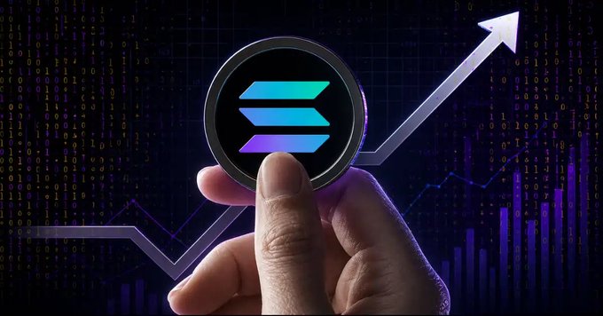 A hand holds up a Solana (SOL) cryptocurrency coin against a dark background featuring code, a rising arrow, and a stock chart.