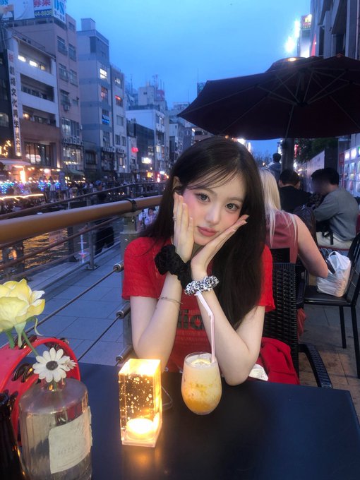 A young woman in a red t-shirt sits at an outdoor cafe table in the evening, resting her chin on her hands next to a drink and a glowing candle, with a busy city street and lit umbrellas in the background.