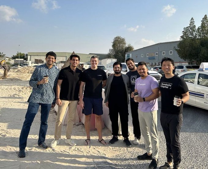 Seven smiling individuals stand together on a sandy construction site, posing for a group photo with industrial buildings, a white police car, and construction equipment visible in the background. One person on the right wears a t-shirt reading "Symbolic Capital," suggesting this is a team outing or social gathering that captures the camaraderie of the "backstreet builders" mentioned in the caption.