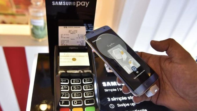 A hand holds a smartphone displaying a Samsung Pay screen near a card payment terminal, with promotional signage reading 'Samsung Pay' and 'Simple. Secure. Works everywhere.'