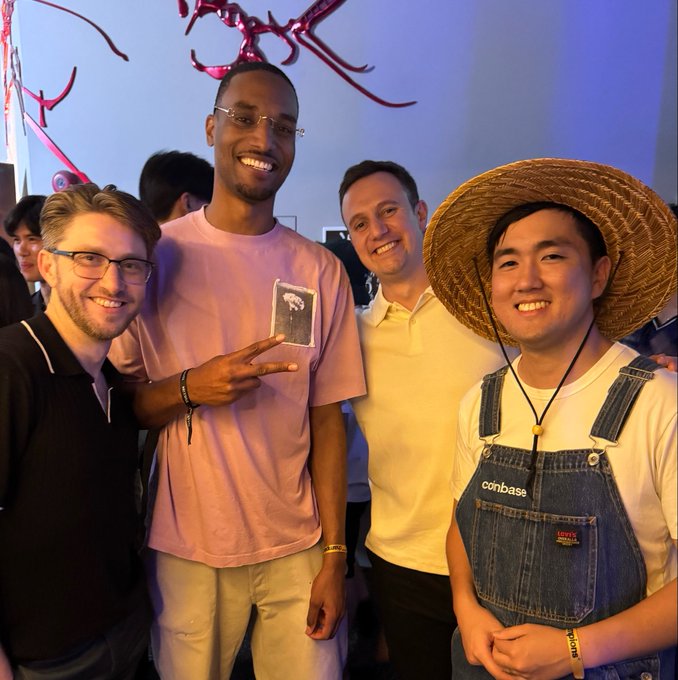 Four men pose together at what appears to be an indoor event with decorative red antlers overhead; one wears a straw hat and a Levi's denim overall with a 'coinbase' logo, while another wears a pink shirt featuring a graphic print.