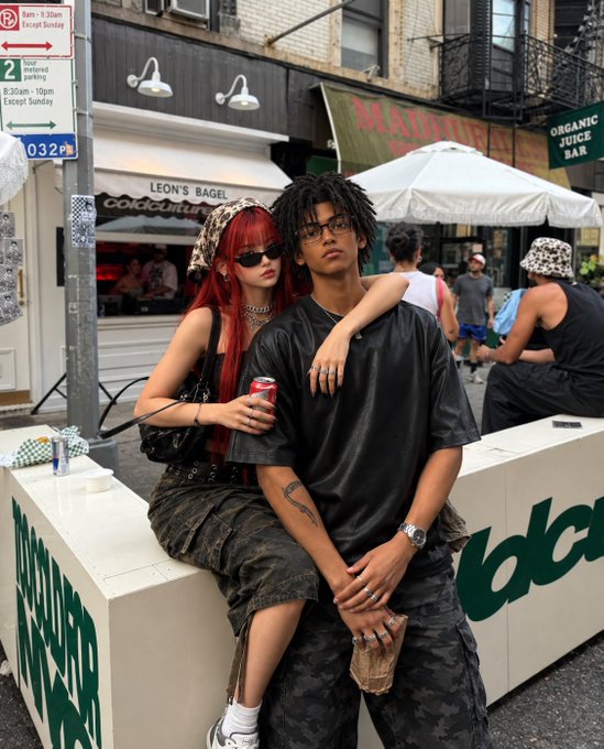 Two young people sitting together on an outdoor bench in front of 'Leon's Bagel' and an 'Organic Juice Bar' in what appears to be New York City, with the woman having red hair and the man wearing black leather attire.