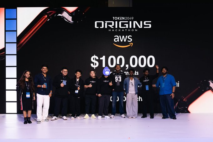 A group of nine people pose onstage beneath a large screen reading “TOKEN2049 ORIGINS HACKATHON,” “aws,” and “$100,000.” Several attendees hold thumbs up, and one person holds a trophy while wearing a purple face covering.